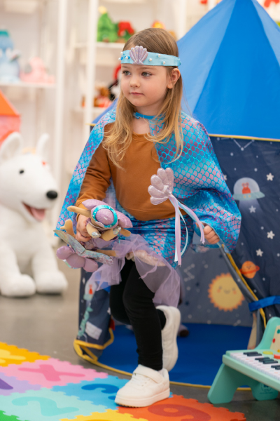 A little girl wearing Gigglescape dress-up clothes in front of a play tent