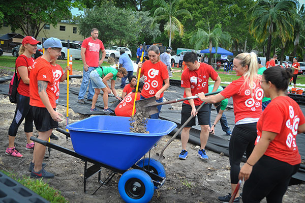 A group of volunteers in red shirts shovel mulch into a wheelbarrow as they build a new play space at a local school.