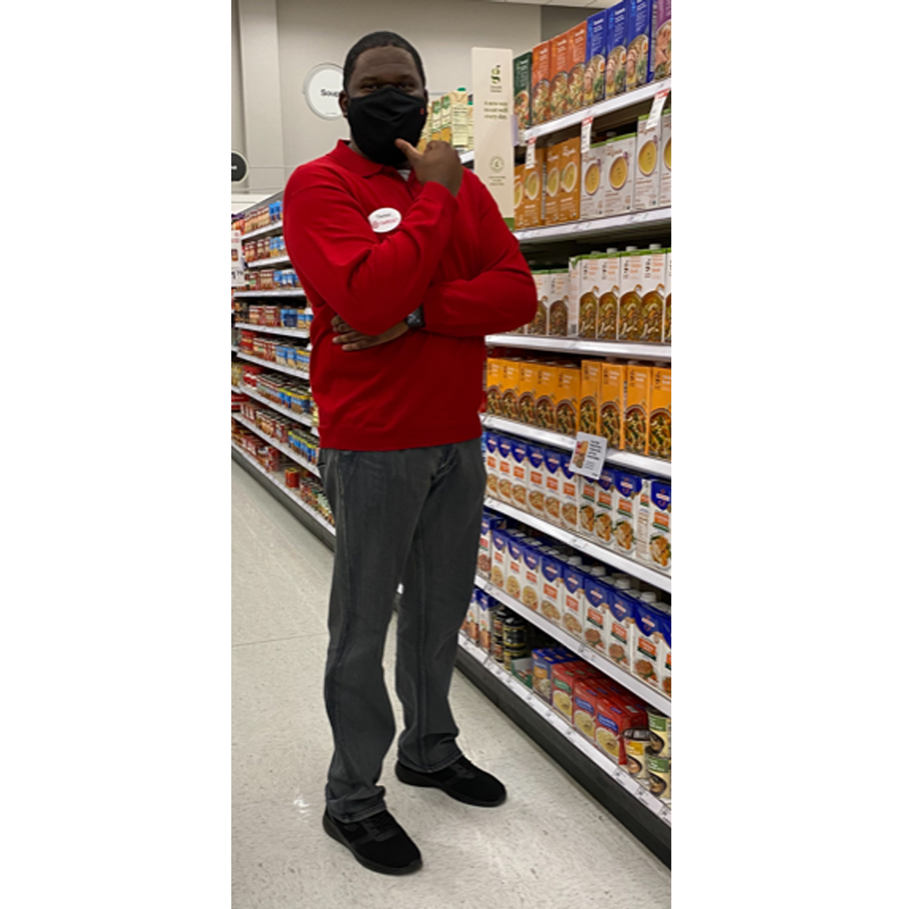 Thomas, wearing a red shirt, name tag and face mask, stands near shelves of soups and canned goods