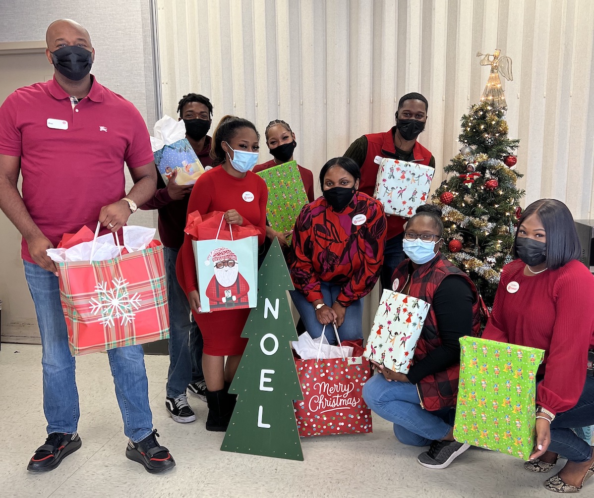 A group of Target team members pose in front of a holiday tree with wrapped and bagged gifts.