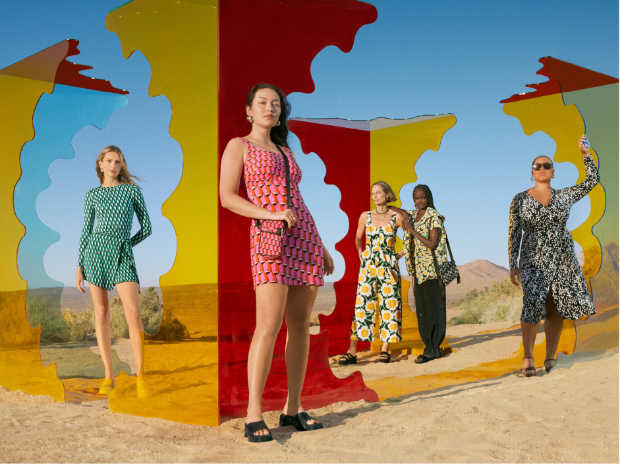 Five women in bright floral dresses, rompers and blouses pose among colorful cutouts in a desert setting.
