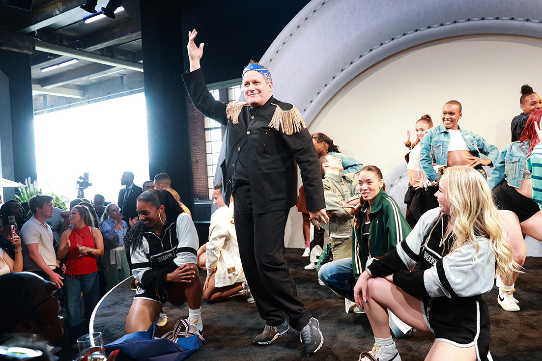 A man stands in a black suit and blue bandana with his arm in the air, smiling at a crowd
