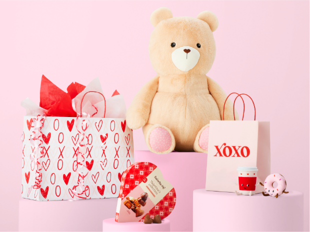 A plush bear, Valentine’s Day gift bags, a box of chocolates and a coffee and donut decoration, all on a pink background.
