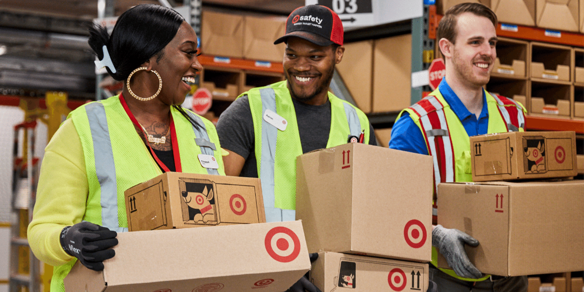 Three smiling Target team members at a supply chain facility carry boxes with Bullseye logos.