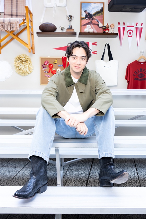 A man sits on bleachers in jeans, cowboy boots and a green jacket