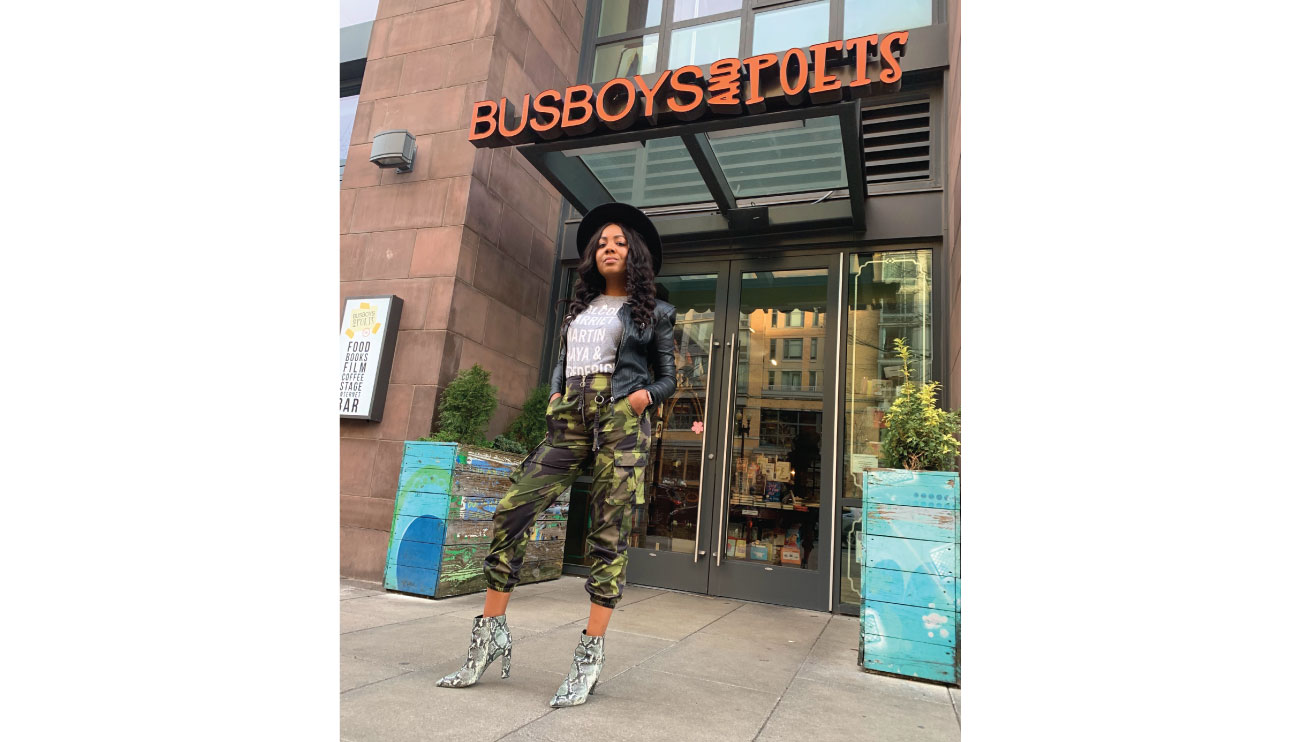 Maya stands in front of a downtown store wearing a white top, black jacket, camo pants, snakeskin high-heel boots and a black hat