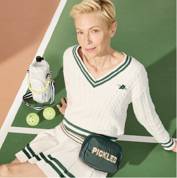 A model sitting on a pickleball court wearing a white sweater and skirt with green trim.