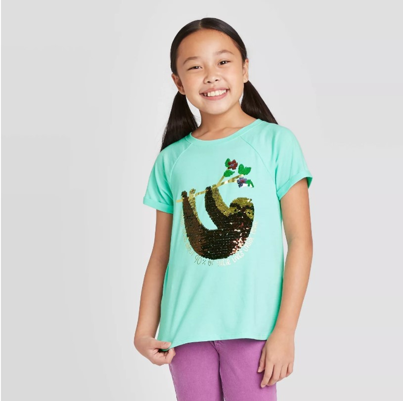 A young girl with pigtails models a mint green tee with a shiny brown and gold sequined sloth hanging from a tree branch.