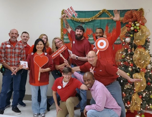 10 smiling team members in red shirts pose together near holiday decor as they hold up work.win.give signs