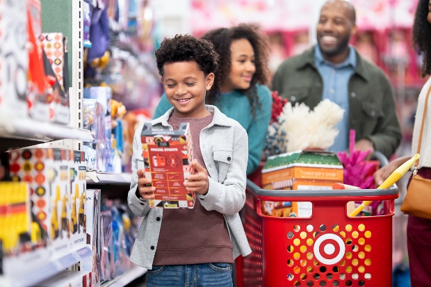 A family is pushing a cart full of merchandise while a child stands in front of the cart holding a toy and smiling.