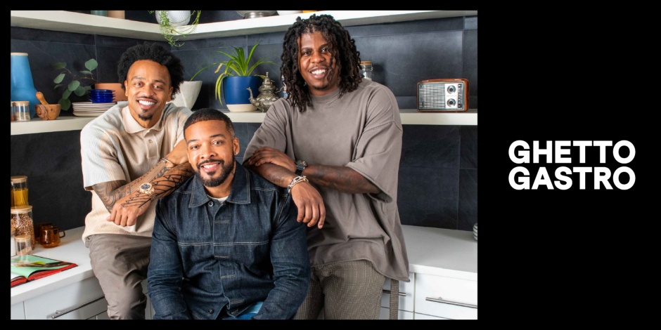 Three people, Ghetto Gastro founders Jon Gray, Lester Walker and Pierre Serrao, sit in a room filled with kitchenware smiling at the camera, next to the Ghetto Gastro logo.