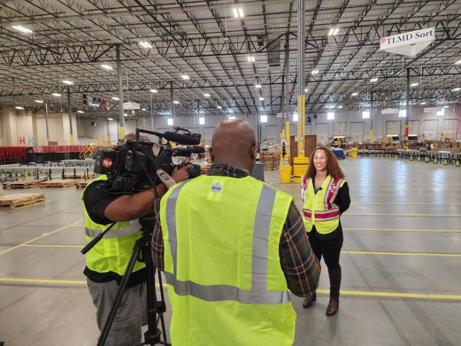 A person stands in front of a news camera, smiling, in the middle of a vast distribution center.