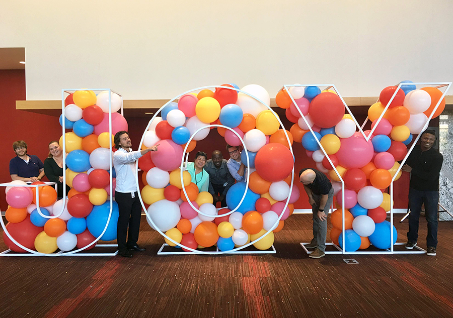 Eight team members pose playfully with a large display of colored balloons that spell out JOY