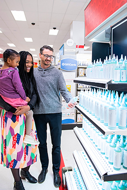 Josef and Camille, who is holding daughter Mila, stand in a Target beauty aisle smiling and looking at their products on a display