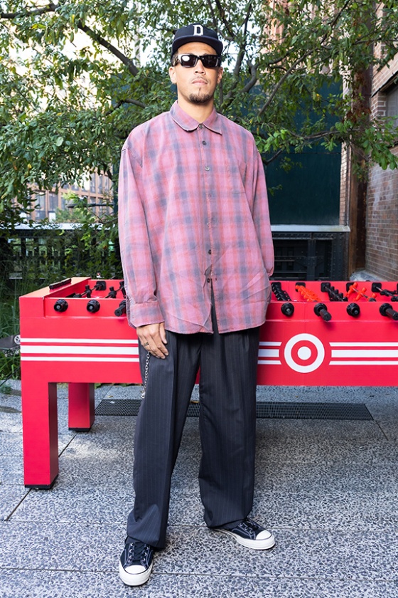 A person stands in front of a foosball table. They wear sunglasses, a hat with the letter “D,” and a plaid shirt.