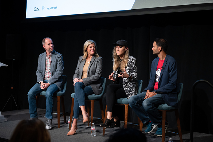 Four panelists, two men and two women, sit on chairs onstage. They are looking at one woman who is speaking.