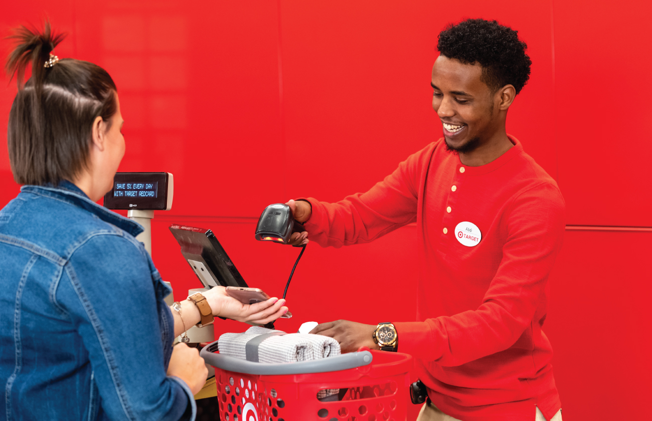 A team member in a red shirt scans a guest's smartphone at the checkout counter against a red wall.