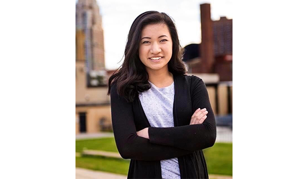 Sophie stands outside in front of a skyline of tall buildings, smiling with arms crossed. She has long, black hair and wears a gray top and black cardigan.