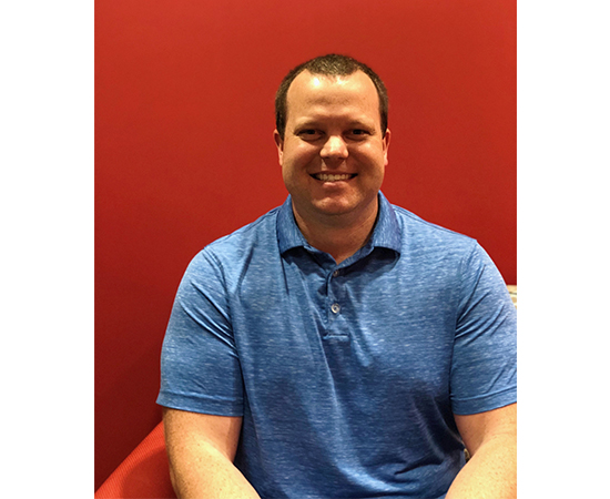 Chad smiles at the camera wearing a blue shirt, sitting against a red background