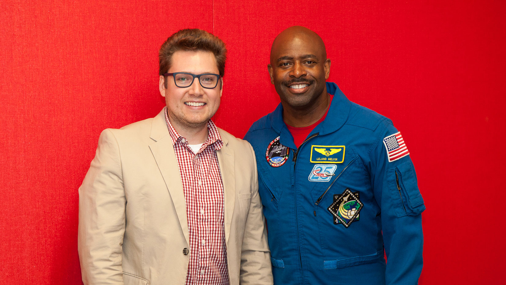 two men stand in front of a red background, one (Leland Melvin) is wearing an astronaut uniform