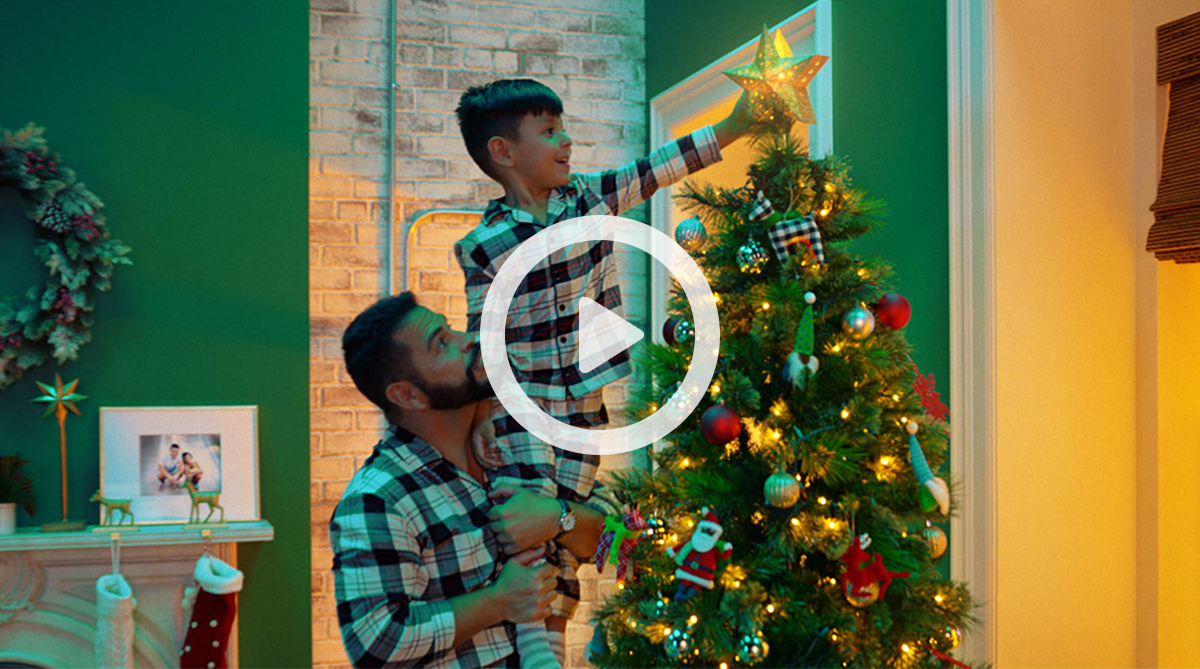 Inside, a man in pajamas holds a young boy in pajamas as he places a star on top of a Christmas tree
