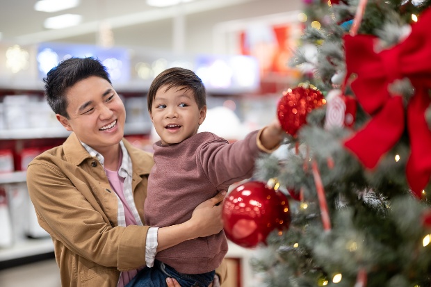 A parent holds a child who is reaching for a red ornament on a tree.