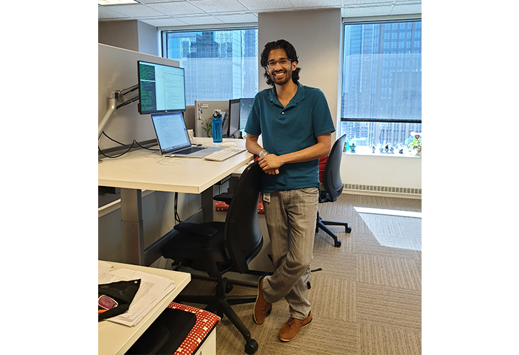Arjun stands in his workspace with his desk and laptop against an office window. He's wearing a blue shirt and tan pants.