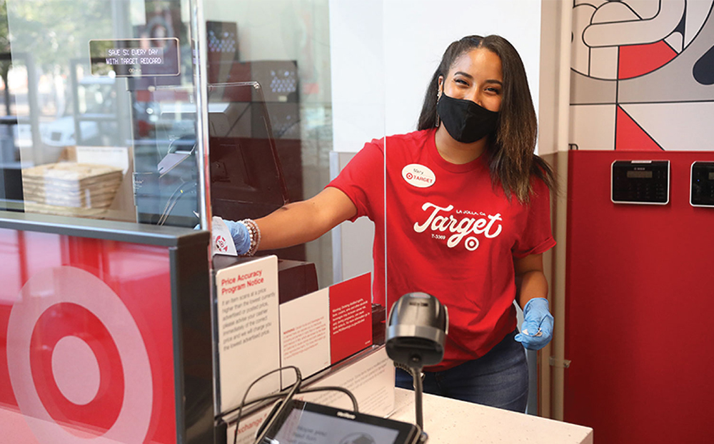 A team member in a red shirt and black face mask smiles from behind a Plexiglas partition as she works the checkout station