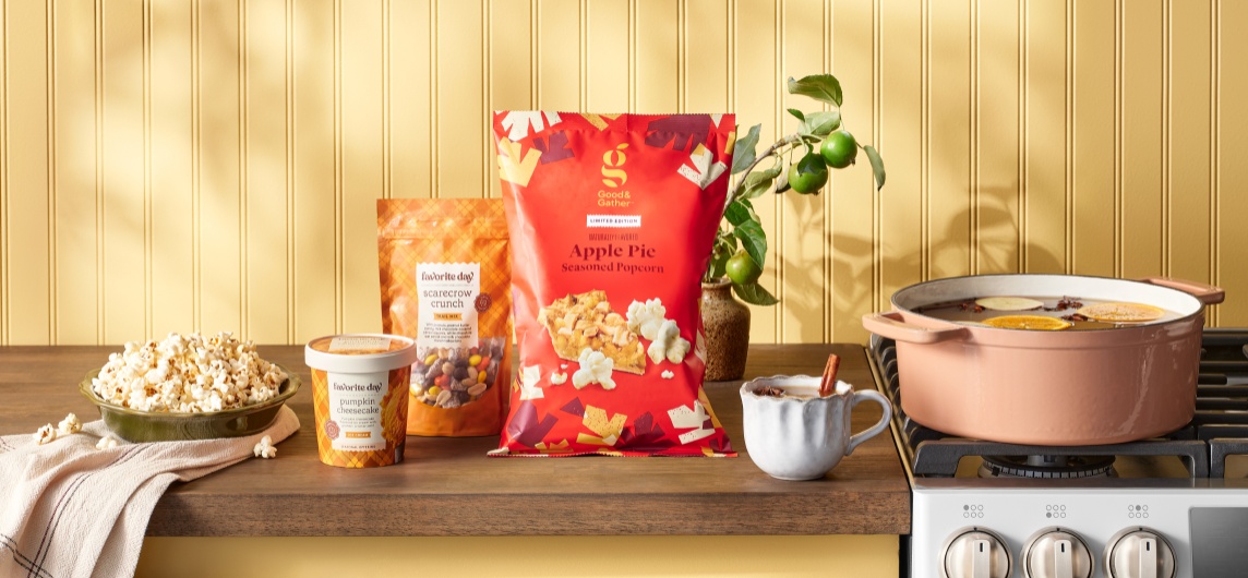 A fall-themed kitchen counter display with Good & Gather Apple Pie Popcorn, Favorite Day Scarecrow Crunch, Pumpkin Cheesecake ice cream, a bowl of popcorn, a mug of cider with cinnamon and a simmering pot of spiced drink on the stove.