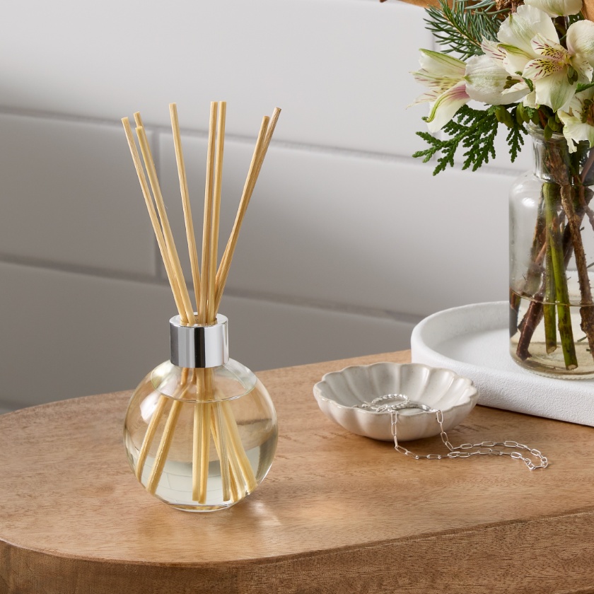 a table with a Threshold reed diffuser next to a small white bowl holding a necklace and a vase of flowers