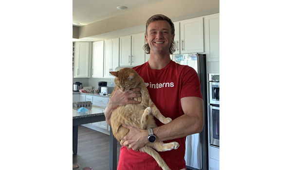 Tres stands smiling in his kitchen at home. He has short blond hair and is wearing a red shirt with white text reading Interns, and is holding his orange cat.