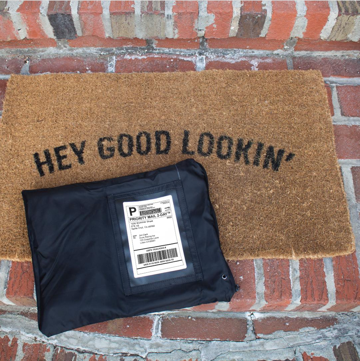 A package sits on a doormat, and the package material is black cloth with a plastic window for the address slip.