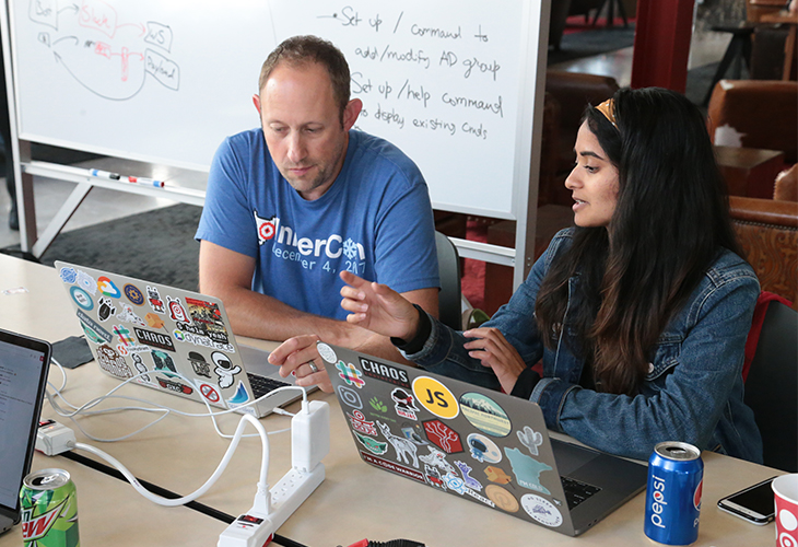 A man and a woman sit at a table with their laptops, which are covered in stickers. There's a whiteboard and chairs behind them.