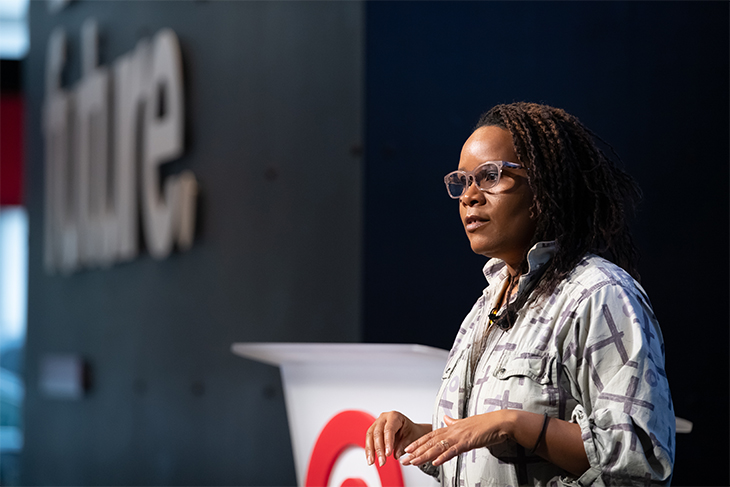 A close-up shot of Niambi onstage, wearing a purple and white shirt, standing in front of the podium, holding up her hands and looking out at the crowd.