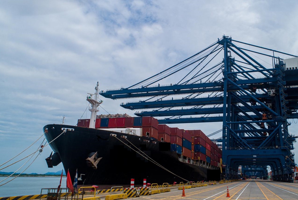 A large ship carrying stacks of shipping containers parked at a dock lined with large cranes for moving freight.