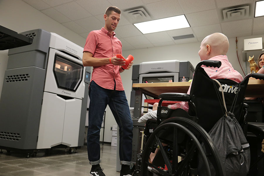 Hallie in her wheelchair looks on as an engineer does a demo in the 3D Design Lab. There are two 3D printers behind him.