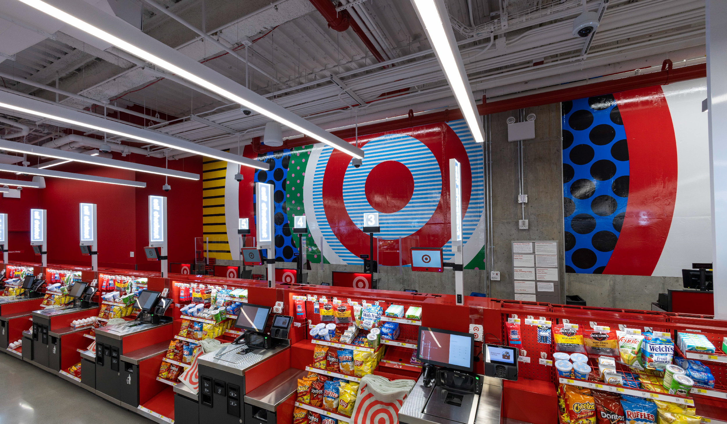 Colorful bullseye artwork hangs on a gray wall behind multiple self-checkout stations.